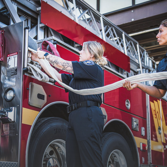 Photograph of two firefighters preparing a firehose in front of a red firetruck with visible large tires and a ladder a woman with tattoos and a smartwatch and another woman with short hair are shown deploying the hose
