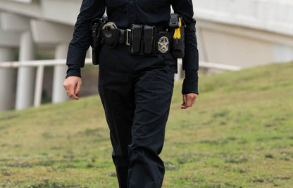 Photograph of a police officer in uniform walking on a grassy hill near a modern bridge with white columns and pillars a gold ring and a yellow utility device are visible on her belt