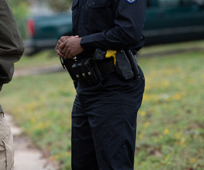 Photograph of a police officer outdoors with a firearm and multiple magazine pouches on their utility belt showing a partially visible dark green vehicle and blurred yellow wildflowers in the background
