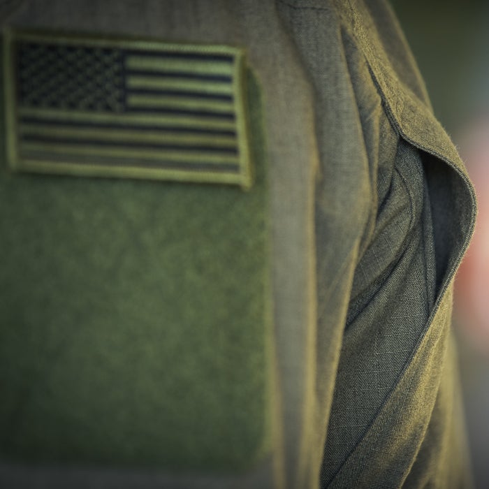 Close-up photograph detail of a military uniform showing an olive-green fabric texture, a US flag patch, and subtle shadowing effects
