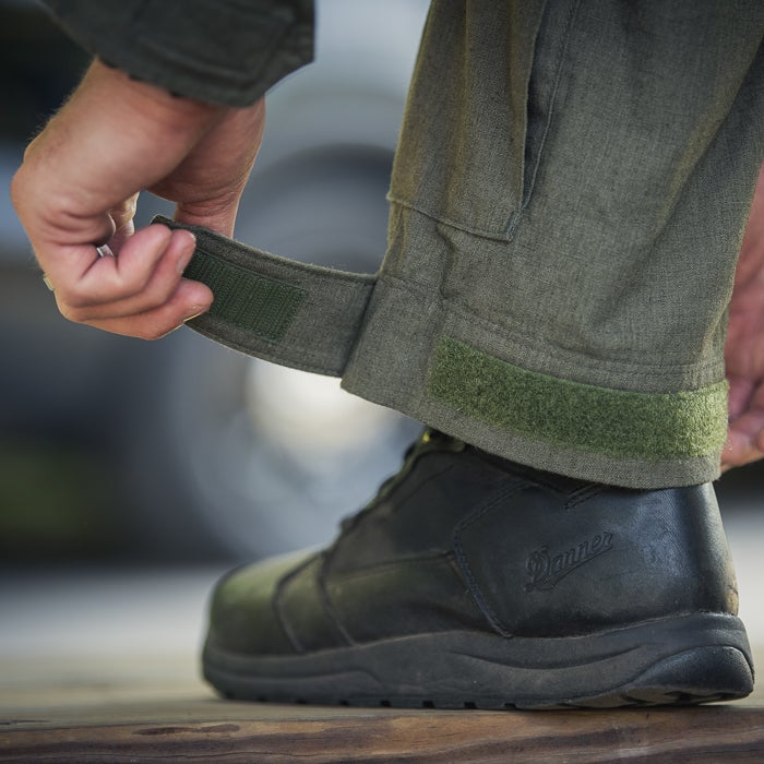 Photograph showing a person adjusting the olive green adjustable cuffs of tactical pants with hook-and-loop closures and wearing black Danner boots with a visible logo on a wooden surface