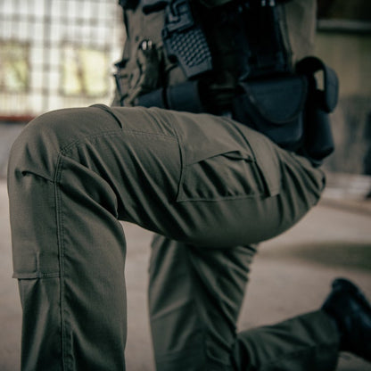 Photograph detail of olive green tactical pants with reinforced stitching and multiple pockets worn by a person kneeling in a derelict building showing textured fabric and dark shadows