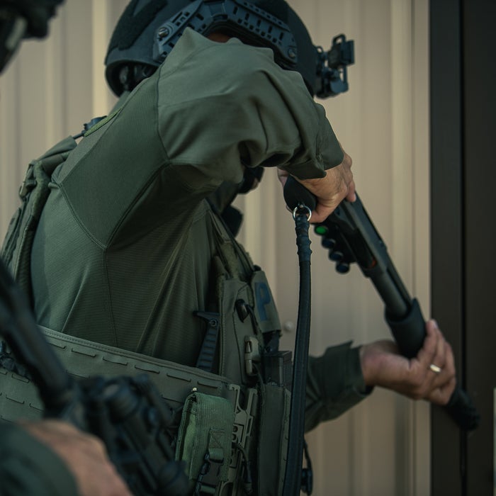 Photograph of a SWAT officer in olive green tactical gear breaching a door with a shotgun a black tactical helmet and vest with pouches are visible
