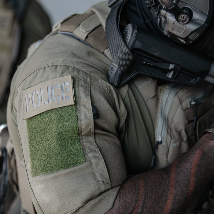 Photograph Police officer’s olive green tactical uniform featuring a POLICE patch, dark green Velcro backing, and a black tactical headset showing a portion of the officer’s arm with visible tattoos.
