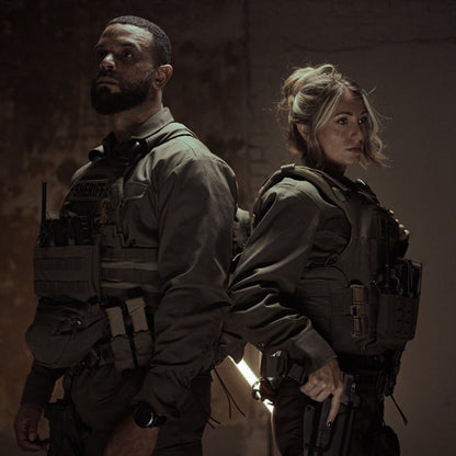Photography: A male and female sheriff stand back-to-back in a dimly lit urban setting wearing tactical vests,  with olive drab uniforms and sidearms visible against a grunge background

