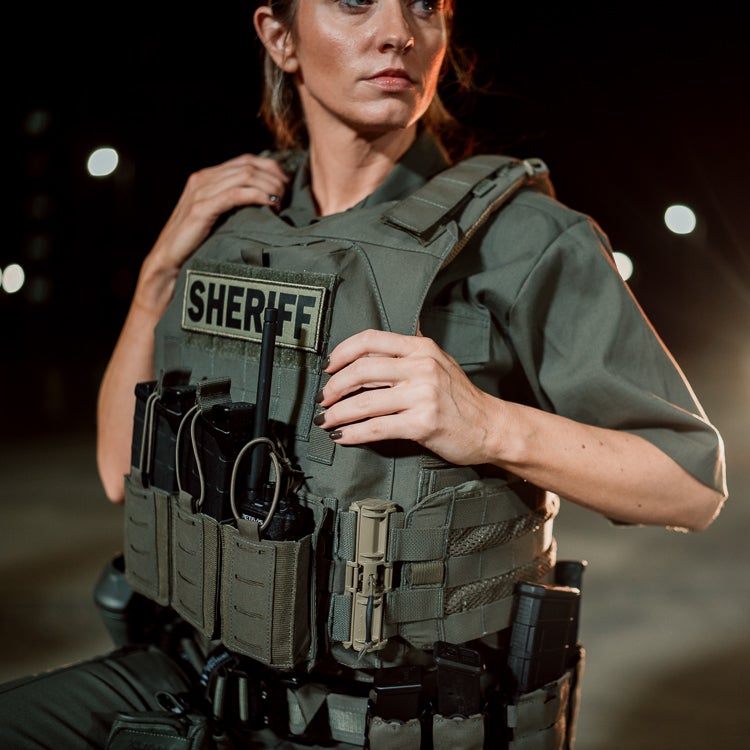 Photography Female Sheriff nighttime outdoor setting  wearing a tactical vest with Sheriff patch and radio dark-toned  olive-green vest and dark nail polish.
