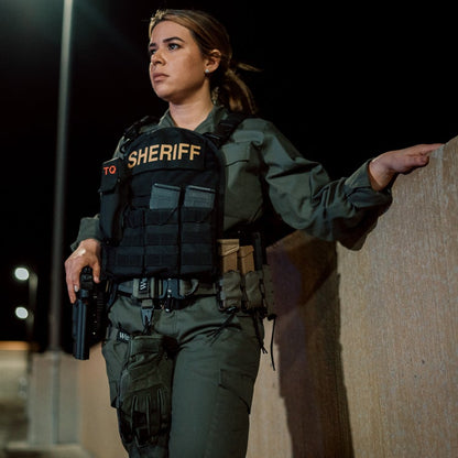 Photography  a female sheriff stands at night against a concrete wall holding a handgun wearing a tactical vest with magazine pouches and olive green pants
