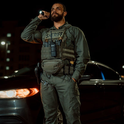 Night photography featuring a Sheriff in olive green uniform with tactical gear and a flashlight, standing before a dark vehicle under city lights at night