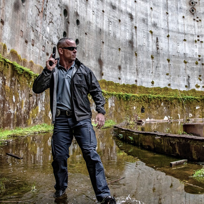 Photography a man in dark tactical gear holding a handgun stands in a flooded abandoned concrete structure with mossy walls and rusted metal debris showcasing his gray shirt and black belt buckle