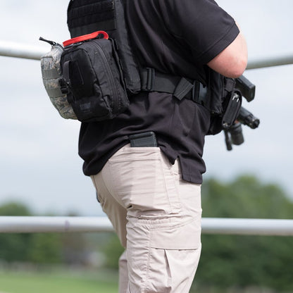 Photograph of a person wearing tactical gear including a black shirt tan pants a black bag with red accents and a rifle magazine in a back pocket outdoors near a metal fence