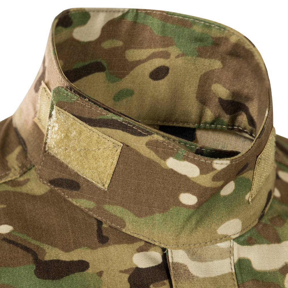 Close-up photograph of camouflage fabric showing a tan velcro patch, detailed stitching, and a blend of green, brown, and beige tones
