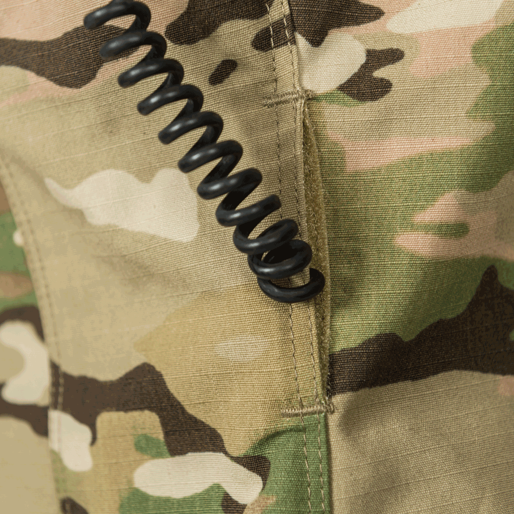 Close-up photograph of camouflage fabric showing a coiled black cord attached to a tan pocket with detailed stitching and a muted green and brown pattern