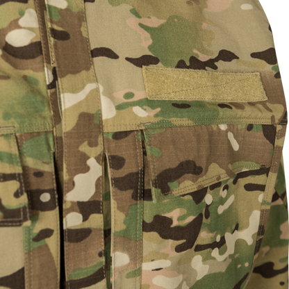 Close-up photograph of OCP camouflage fabric showing a tan nametape and chest pockets with subtle stitching details and a multi-toned pattern of greens, browns, and tans.
